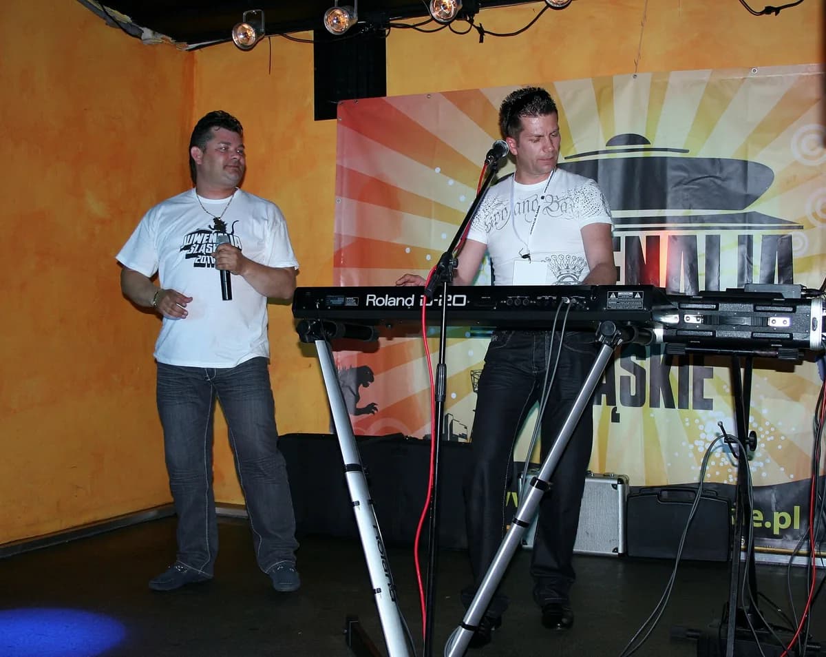 Zenon Martyniuk singing into a microphone while Ryszard Warot plays keyboard beside him, performing on a small stage decorated with a colorful festival banner