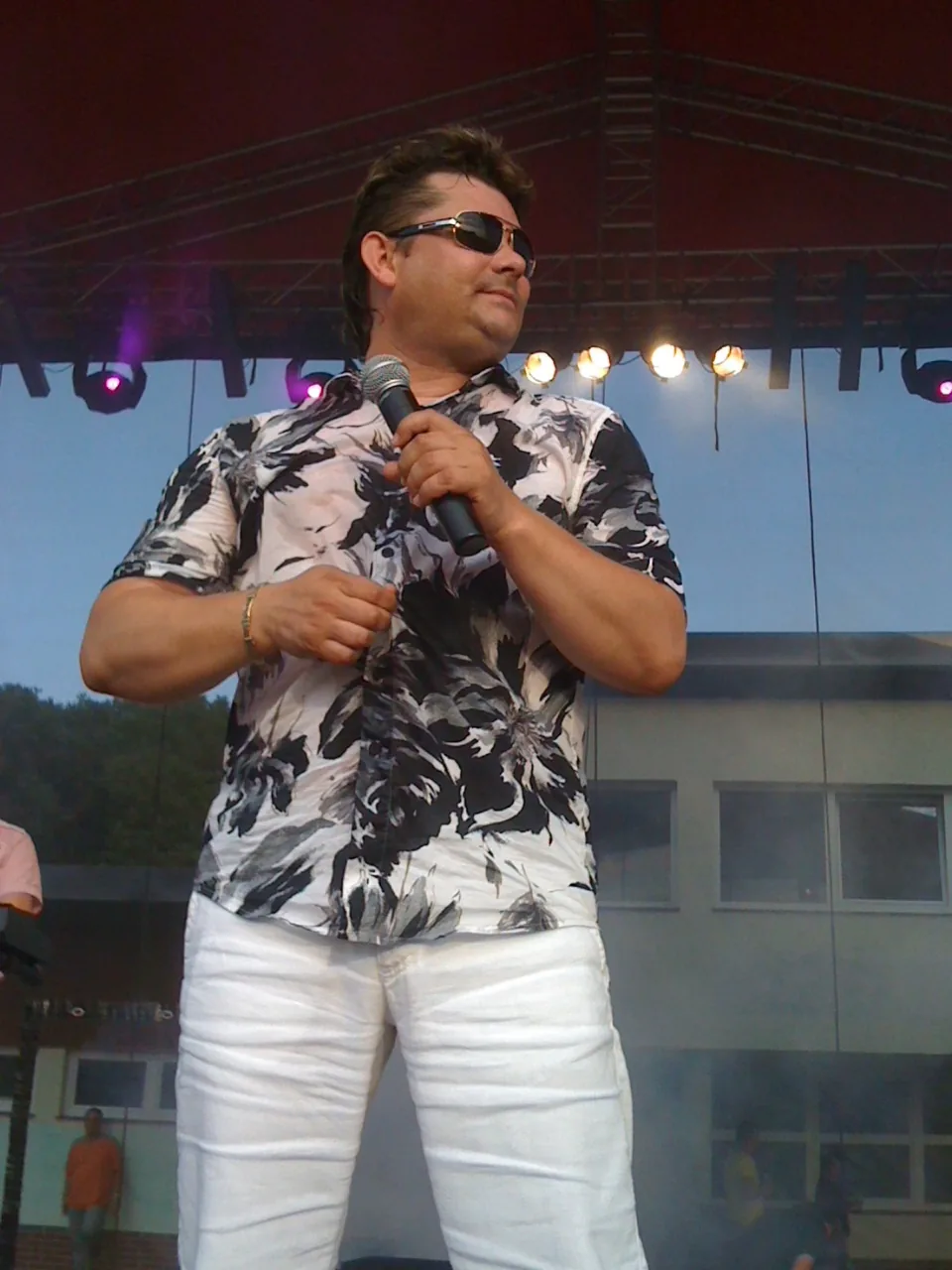 Zenon Martyniuk performing on an outdoor festival stage, wearing sunglasses and a patterned shirt, holding a microphone