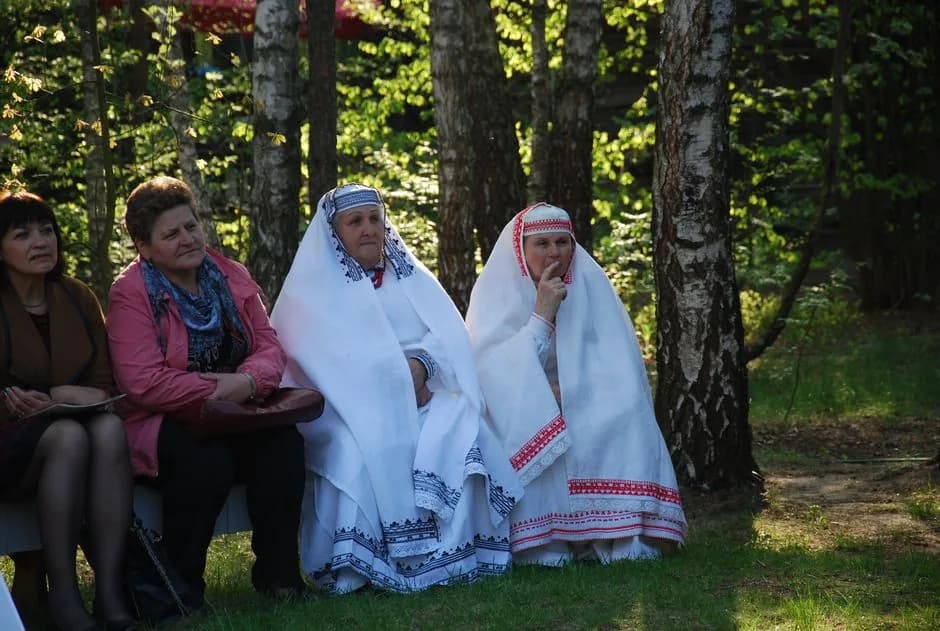 Two women dressed in white ceremonial folk costume with red and black embroidered trim, seated outdoors under birch trees