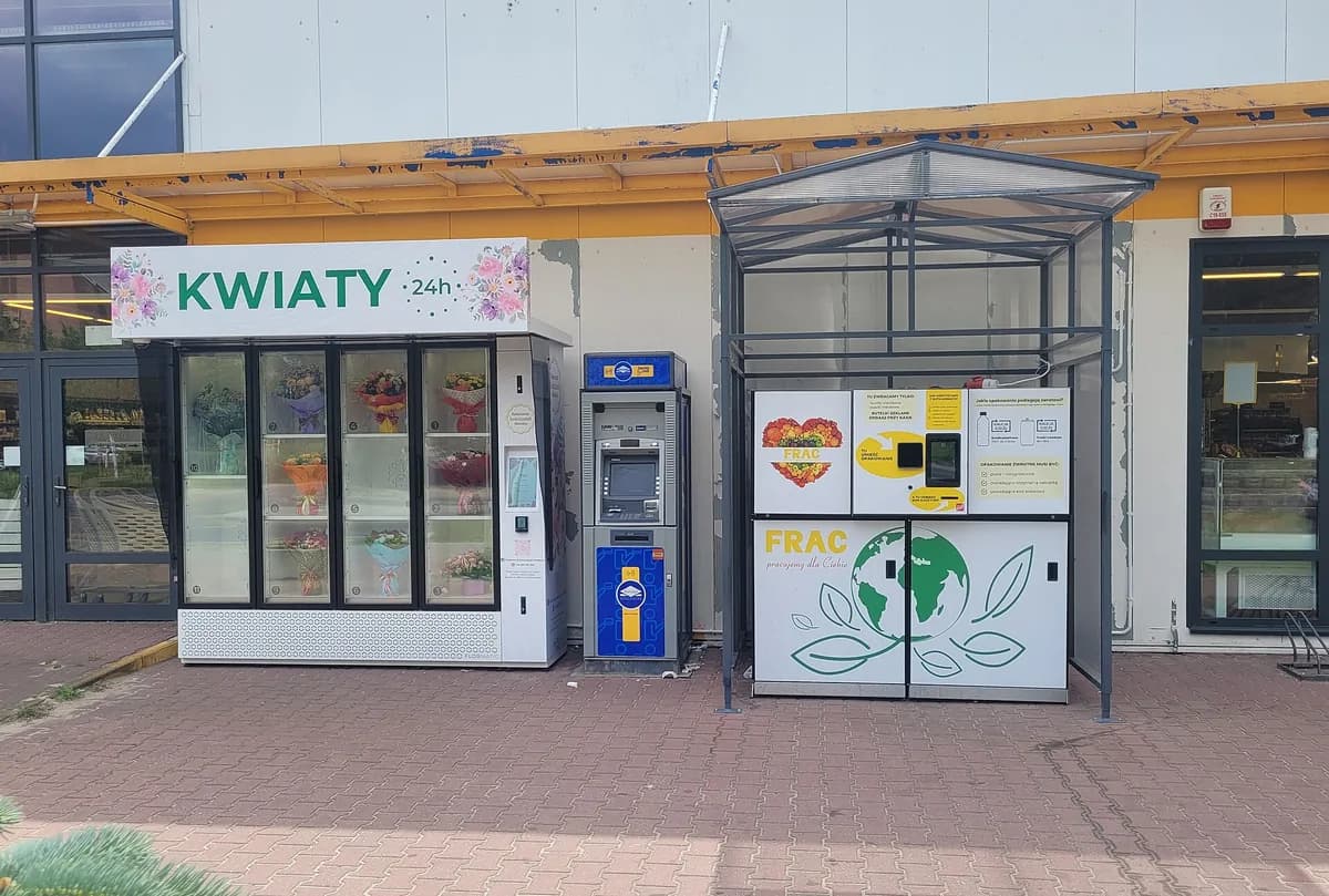 A Kwiaty 24h automated flower vending machine outside a store, its glass compartments stocked with wrapped bouquets, next to an ATM and a recycling machine