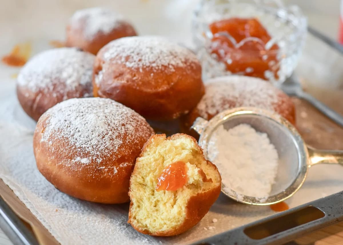 A tray of powdered-sugar-dusted paczki donuts with jam and a sugar sifter