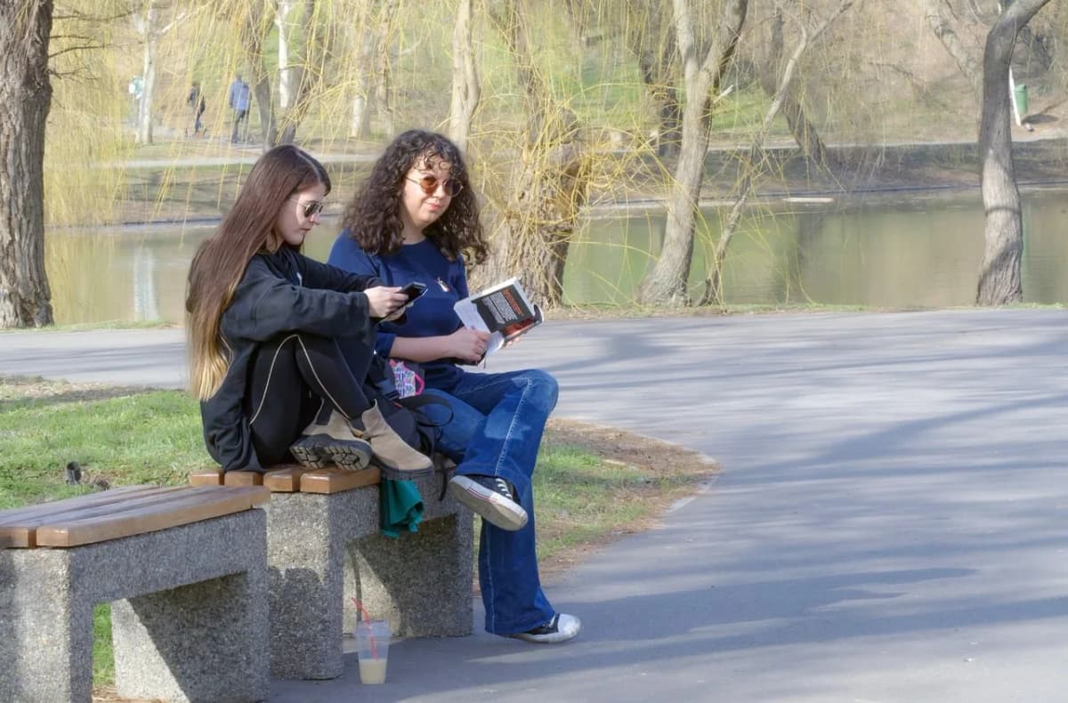 Two friends sitting on a park bench, one checking her phone