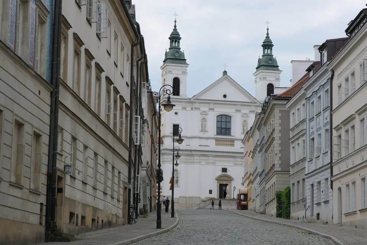 A cobblestone street in Warsaw's Old Town leading toward a white baroque church with twin towers, framed by townhouses on either side