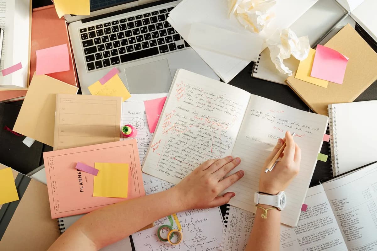 An overhead view of a cluttered study desk with an open notebook full of handwritten corrections, sticky notes, a laptop, and a planner