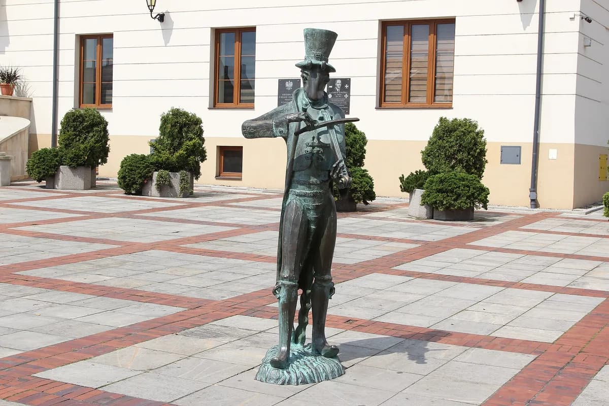 A wide view of the same beetle statue standing in the paved market square of Szczebrzeszyn, with surrounding town buildings visible in the background