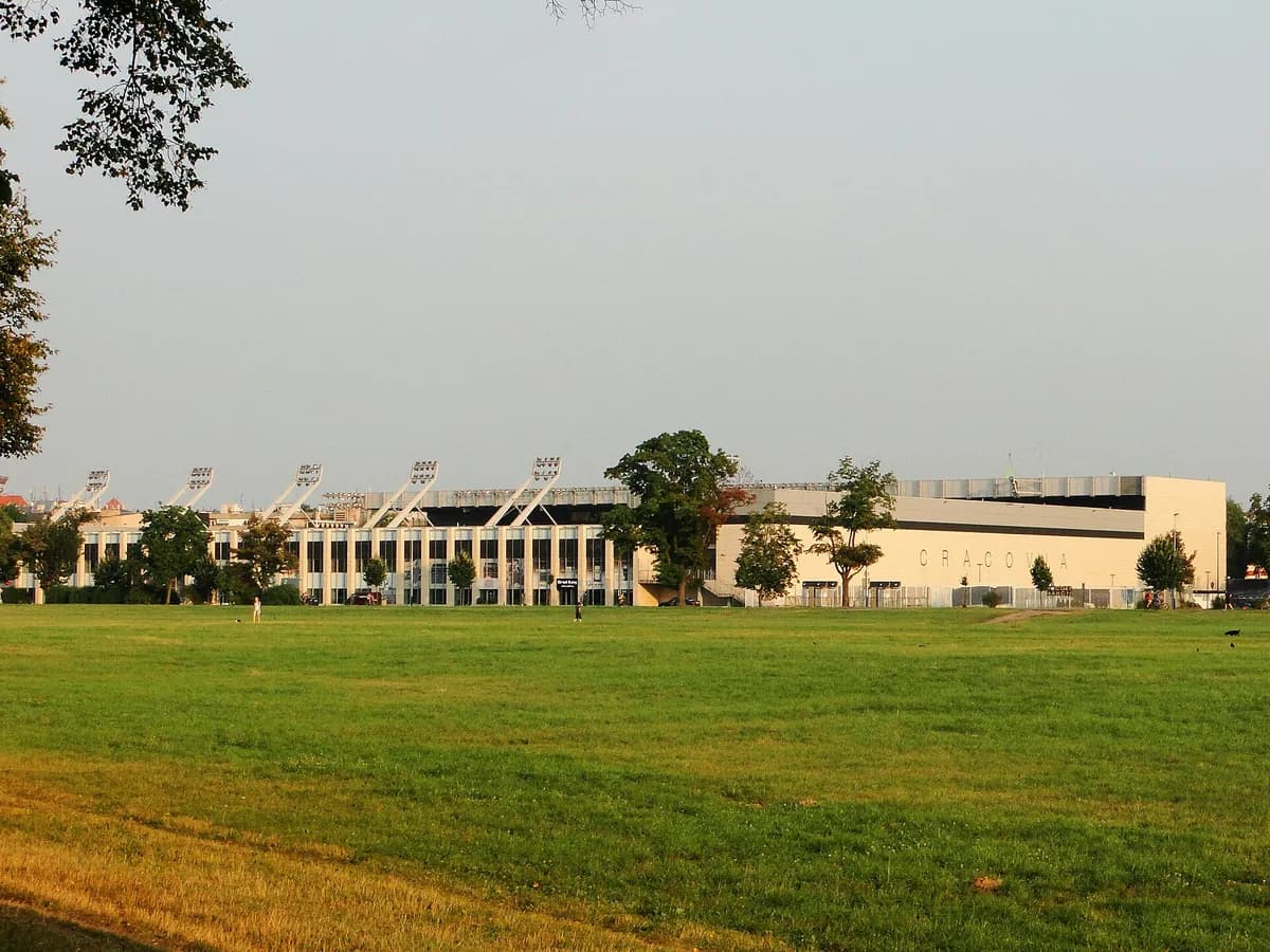 The exterior of Cracovia's stadium in Kraków, a modern building with angled floodlight masts along the roofline, seen from the adjacent park