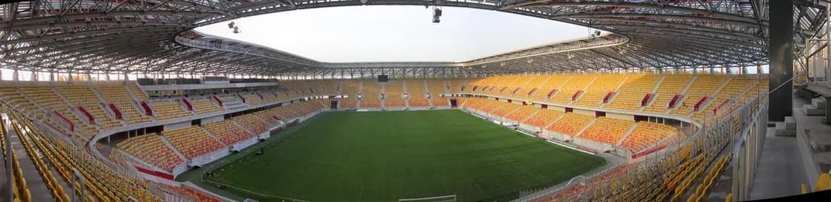 A panoramic interior view of Jagiellonia Białystok's stadium, showing orange and red seating around an empty pitch under a covered roof