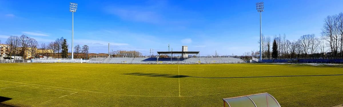 The modest home stadium of Raków Częstochowa, showing simple seating stands, floodlights, and an artificial pitch under a clear blue sky