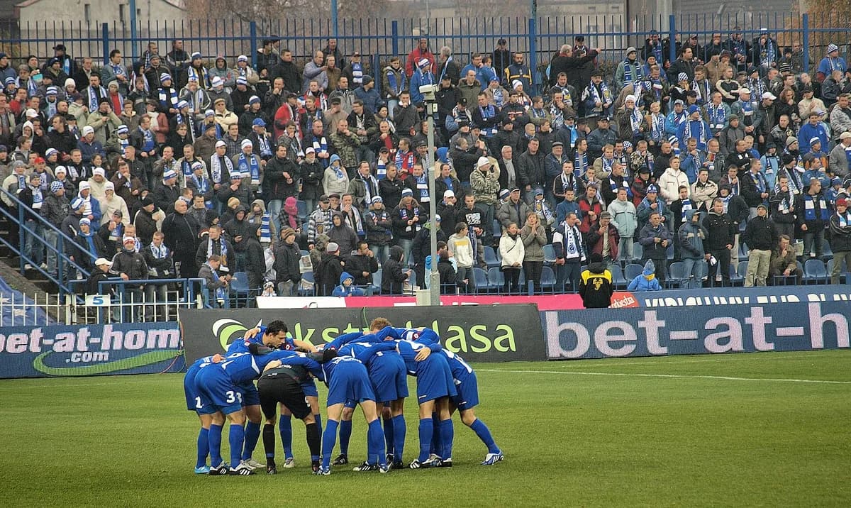 Ruch Chorzów players huddling on the pitch in front of a packed, scarf-waving crowd at their home stadium