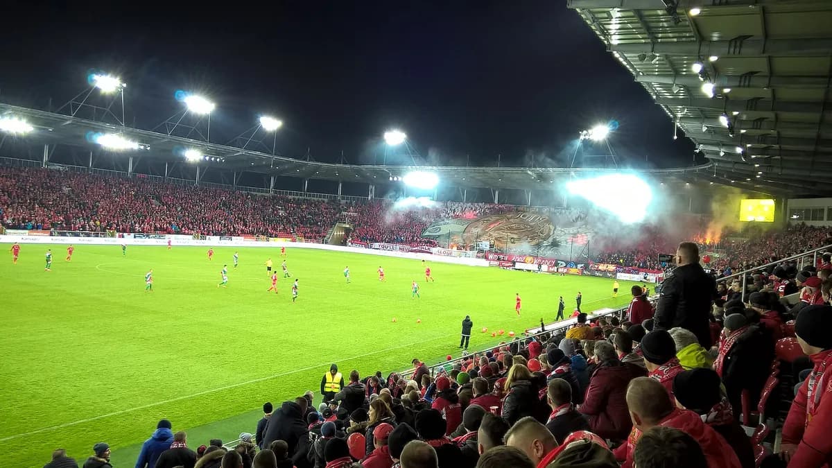 The interior of Widzew Łódź's modern stadium at night, packed with fans in red and white, with smoke and a large tifo display behind one goal