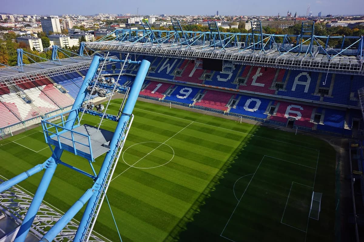 An aerial view inside Wisła Kraków's stadium, its red and blue seats spelling "WISLA 1906," with a TV camera crane in the foreground and the Kraków skyline behind
