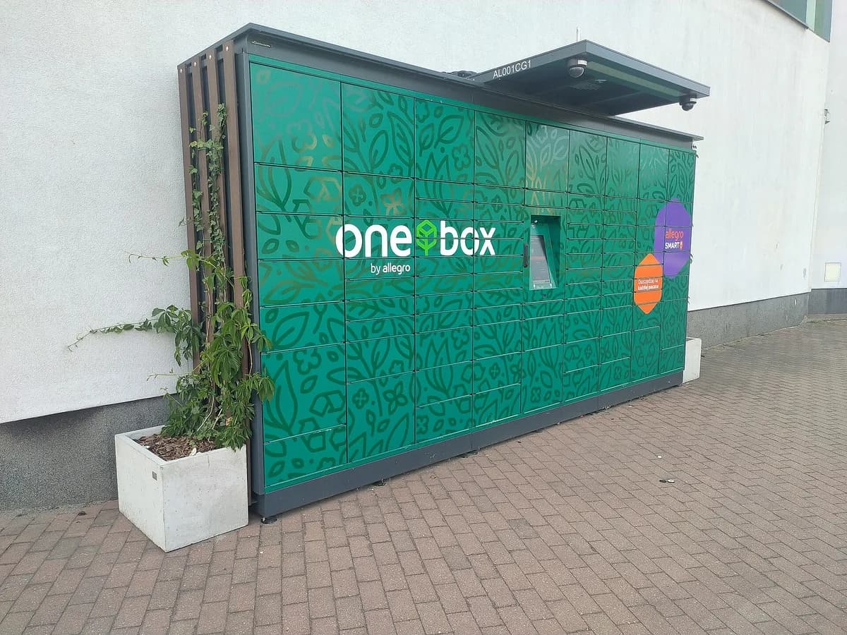 A close-up view of a green "One Box by allegro" parcel locker installed against a building wall, showing the touchscreen interface and an "allegro SMART" promotional panel
