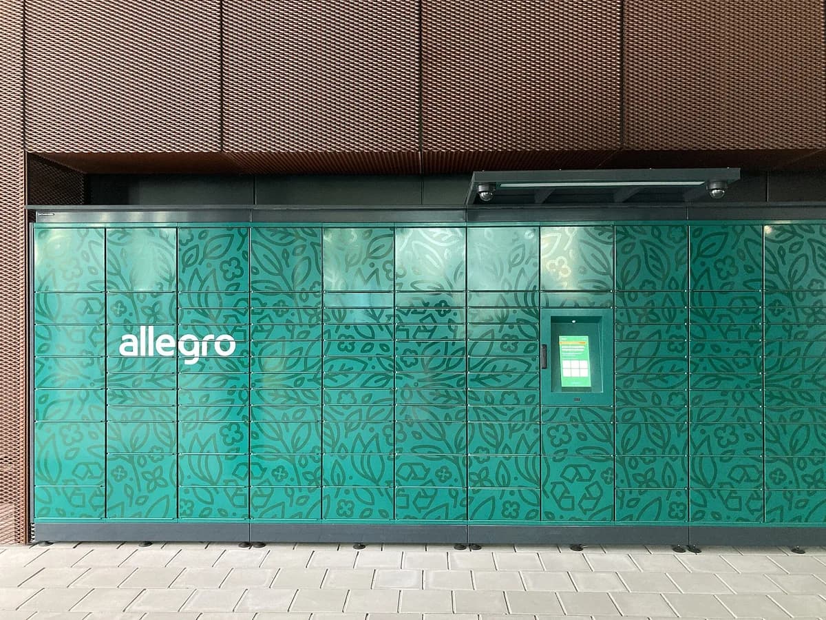 A wide street-level view of a green Allegro-branded parcel locker installation, marked with the Allegro logo, standing against a building facade in Poznań