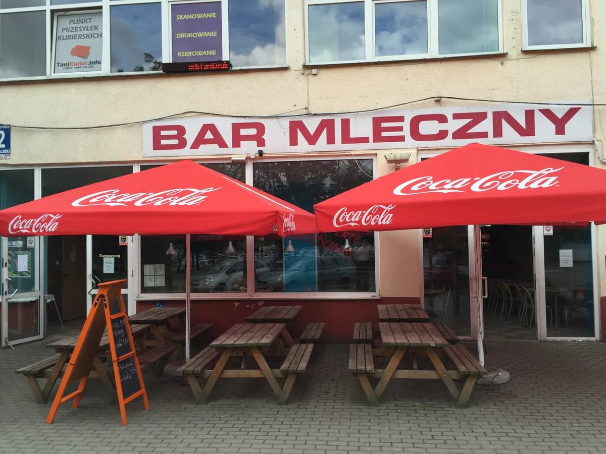The exterior of a bar mleczny in Gdańsk, with outdoor picnic tables shaded by red Coca-Cola branded umbrellas and a "BAR MLECZNY" sign above the entrance