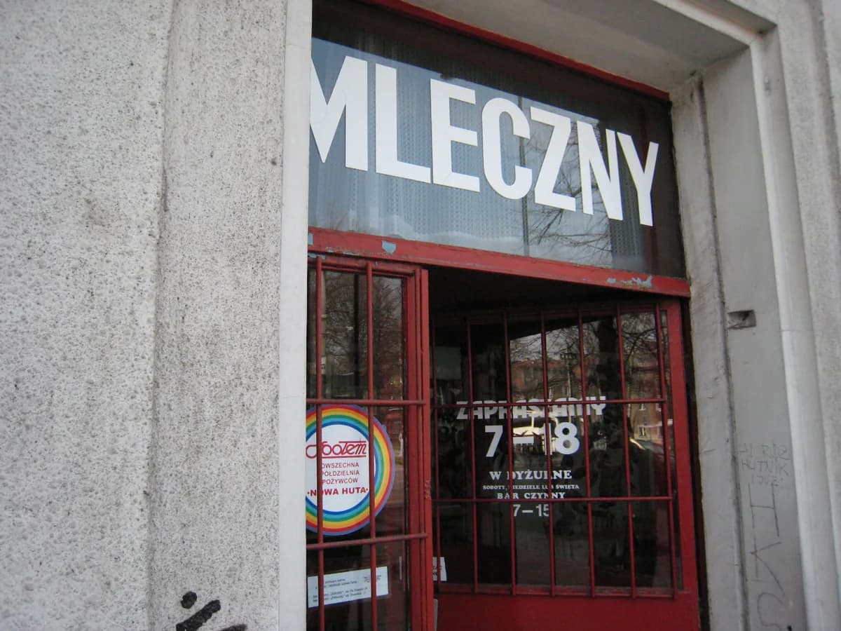 A close-up photo of a "MLECZNY" sign above a red-framed doorway in the Nowa Huta district of Kraków, with a rainbow-colored cooperative society sticker visible on the glass