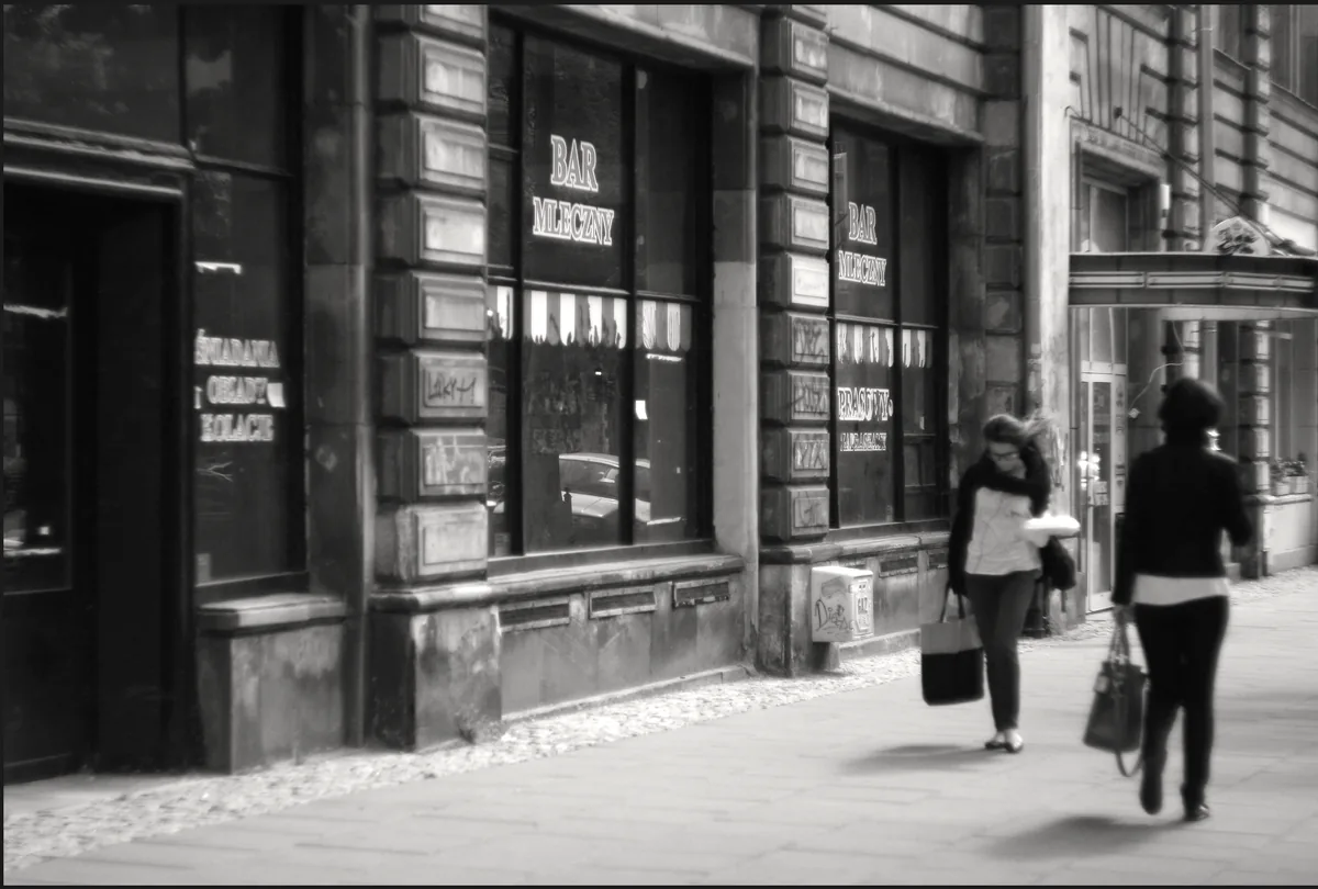 A black-and-white street photograph showing two adjacent storefronts with "BAR MLECZNY" signage, including one reading "Prasowy," with pedestrians walking past on a European city sidewalk