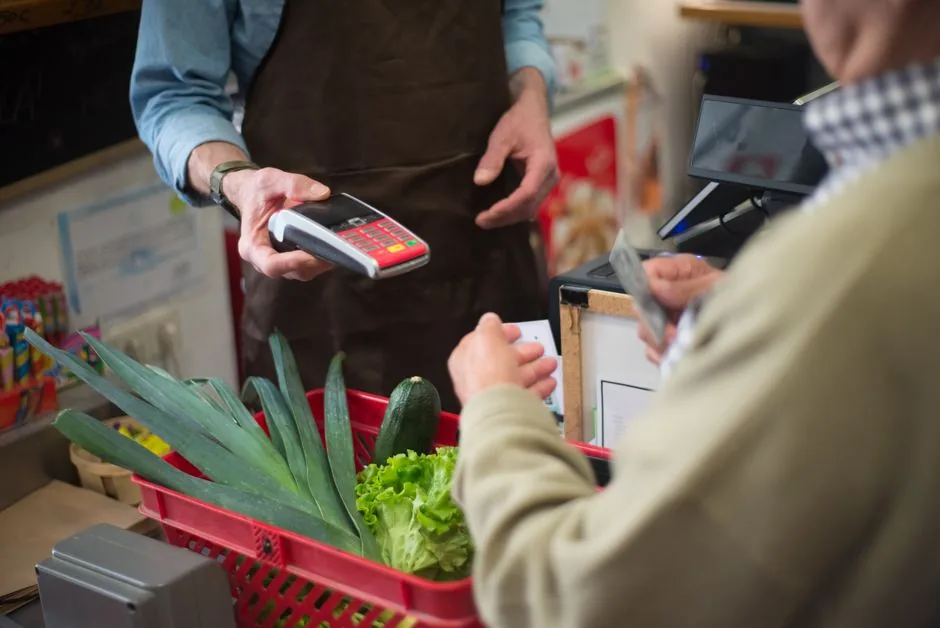A customer paying by card at a small grocery checkout counter, with a basket of fresh vegetables on the counter