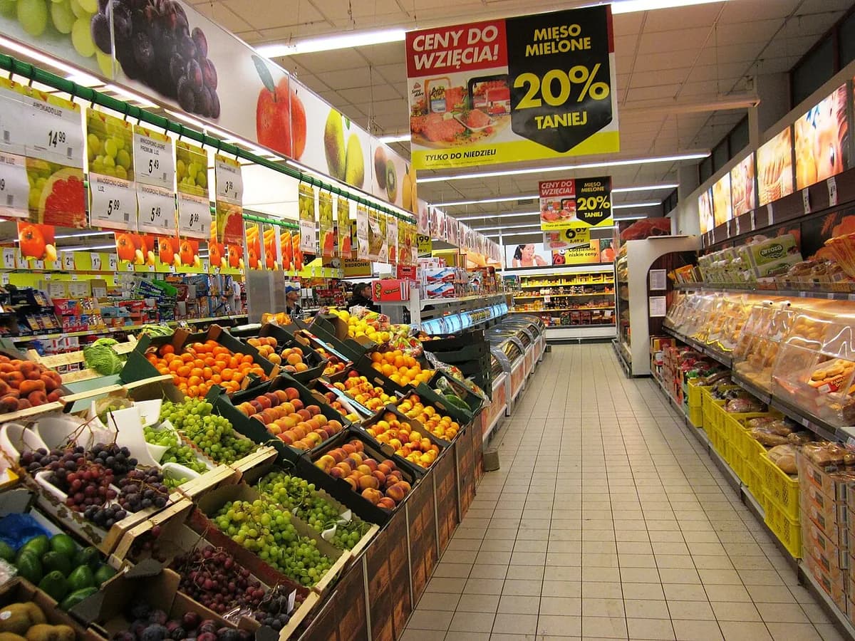 The interior of a Biedronka supermarket showing a full produce aisle with fresh fruit, a bakery section, and promotional signage advertising discounted meat