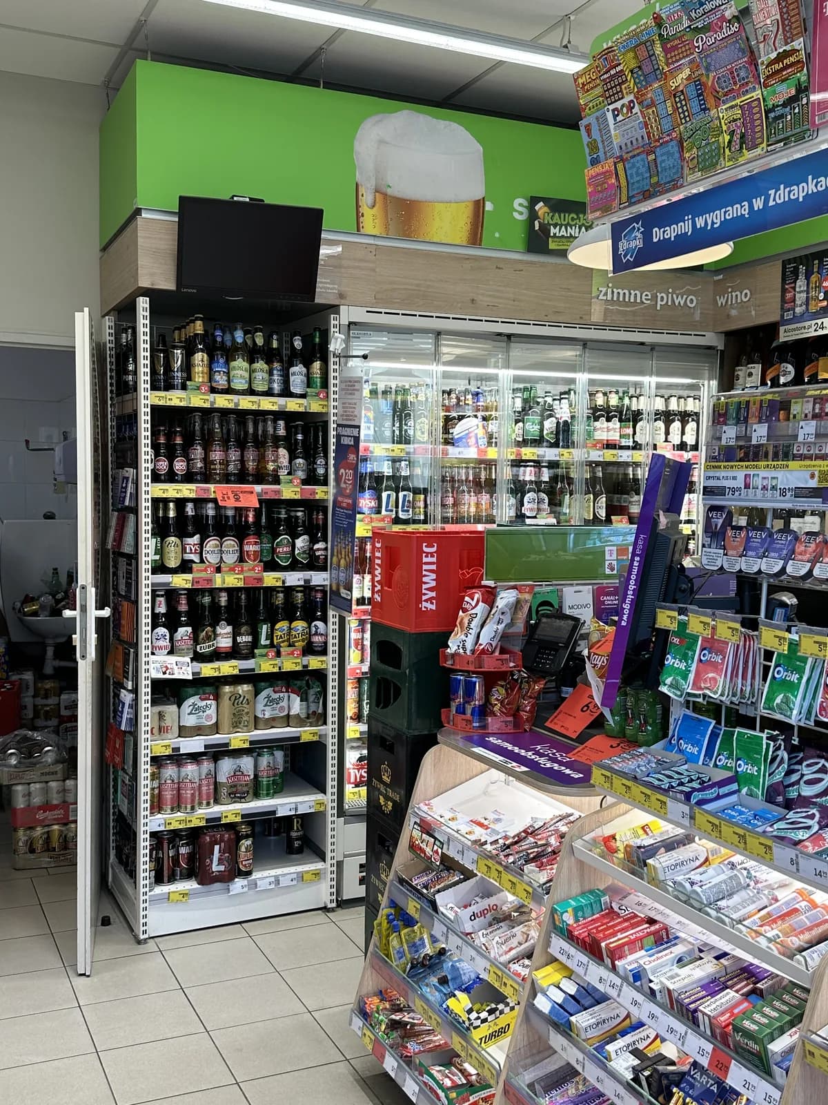A glass-door cooler stocked with rows of Polish and international beer bottles and cans inside a Żabka