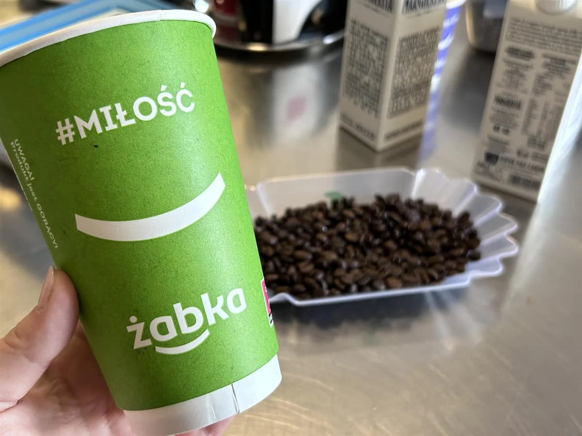 A Żabka Café paper cup printed with the frog logo and the hashtag #miłość, held next to fresh coffee beans at the in-store counter