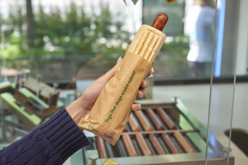 A Żabka Nano hot dog wrapped in its branded paper sleeve, fresh off the automated grill inside a store