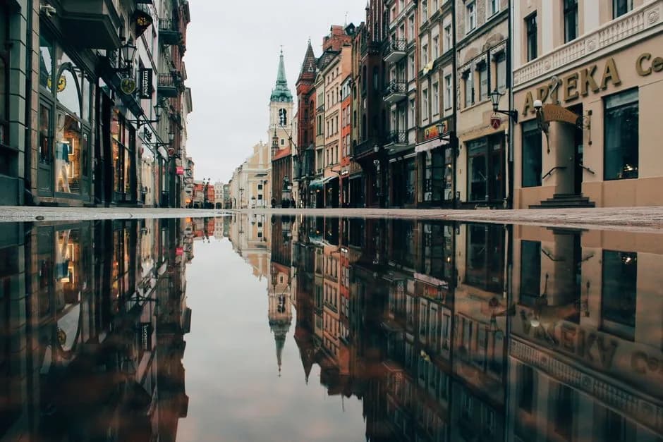 A quiet European street at dusk with shopfronts and reflections in a wet pavement, the kind of everyday corner where a Żabka would sit