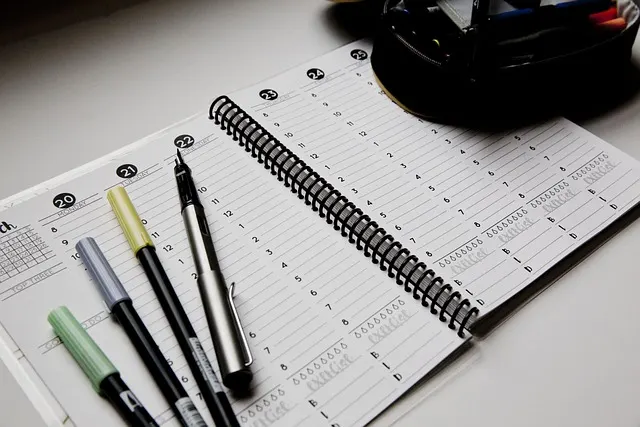 A spiral-bound desk planner open to a weekly grid, with several colored pens resting on top