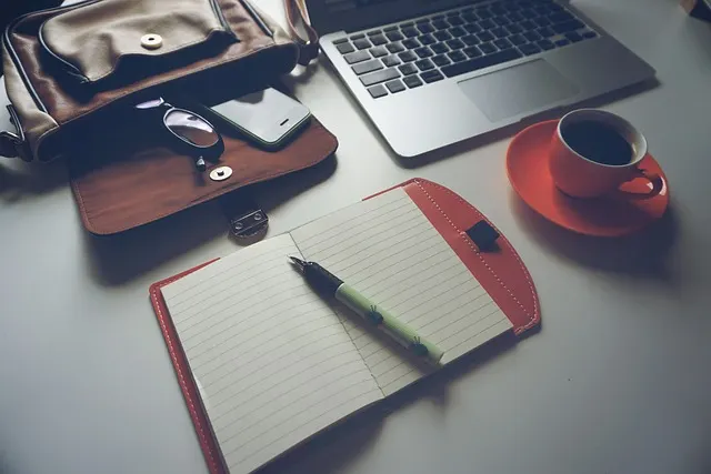 A desk flatlay with an open laptop next to a physical notebook, pen, and coffee — a typical setup for practicing Polish writing