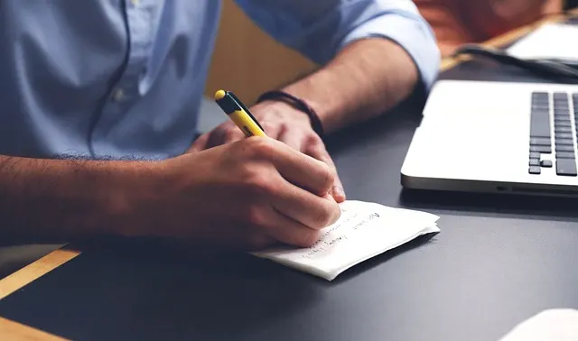 A person writing handwritten notes on paper next to an open laptop