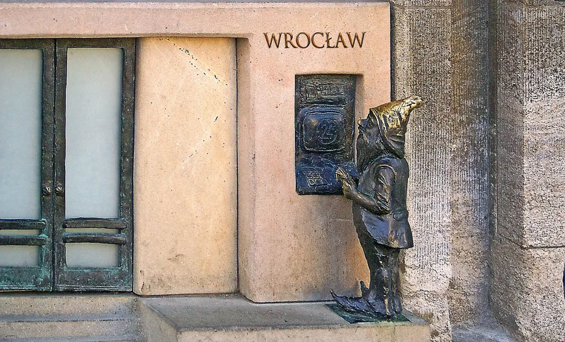 A small bronze krasnal gnome statue beside a doorway intercom in Wroclaw
