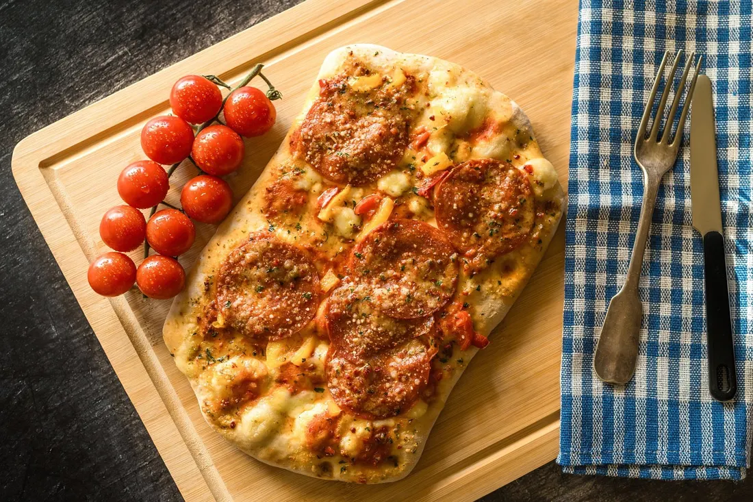 A rectangular baked flatbread topped with melted cheese, spiced sausage, and cherry tomatoes on a wooden board, similar to modern gourmet takes on zapiekanka